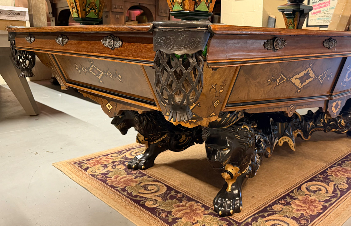 Fully restored antique wood billiard table featuring hand-carved black lion pedestal legs, detailed wood inlay, and traditional leather net pockets.